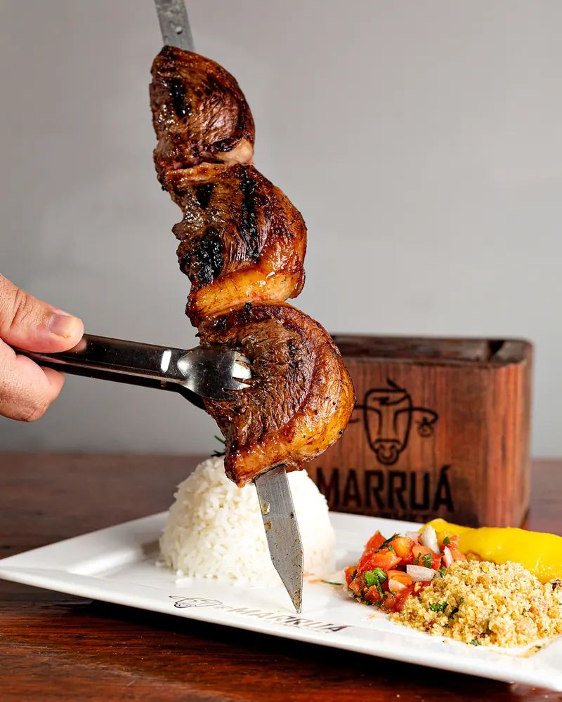 Espeto de carne sendo servido à mesa com pegador, ao lado de arroz, vinagrete e farofa