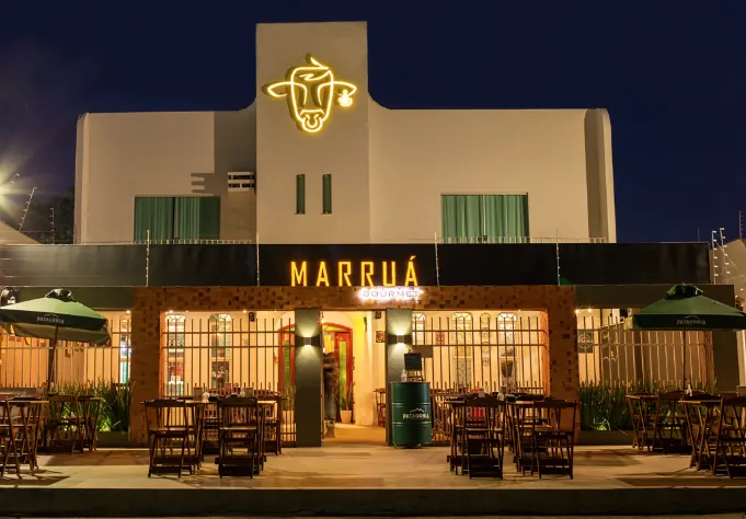 Fachada da Marruá Gourmet à noite: casarão claro com a cabeça de boi em neon âmbar e mesas na área externa