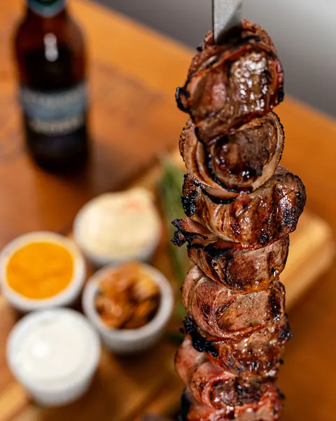 Espeto de carne saído da brasa, com potinhos de molho ao fundo