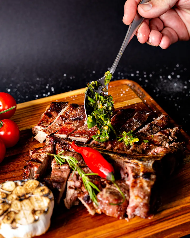 Corte bovino na brasa fatiado sobre tábua de madeira, recebendo chimichurri, com pimenta dedo-de-moça e alho assado